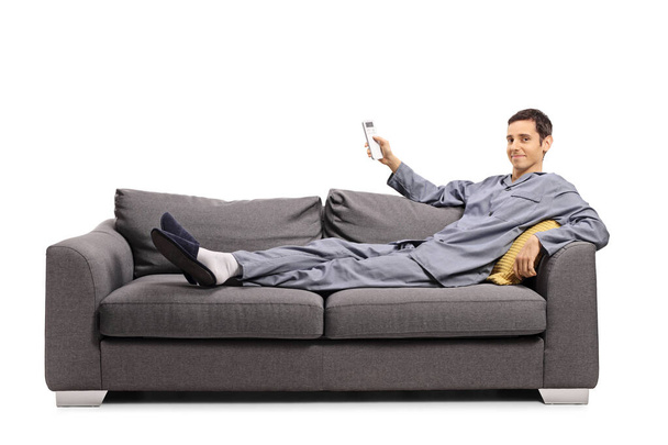 Man in pajamas laying on a sofa and holding a remote control from an air conditioning isolated on white background - Photo, image
