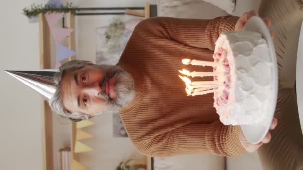 Vertical medium point-of-view shot of caucasian middle-aged man in silver cap celebrating birthday via video chat smiling while blowing out candles on birthday cake - Footage, Video