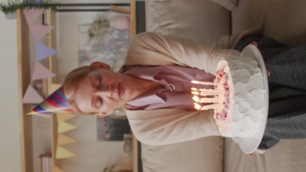 Imágenes verticales de punto de vista medio de una atractiva mujer caucásica de mediana edad con gorra festiva sentada en el sofá con pastel de cumpleaños soplando velas celebrando en casa mediante videollamada - Imágenes, Vídeo