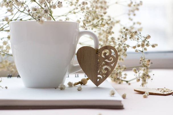 dřevěné srdce a bílý porcelán pohár s kyticí malých bílých květin na světlém pozadí - Fotografie, Obrázek