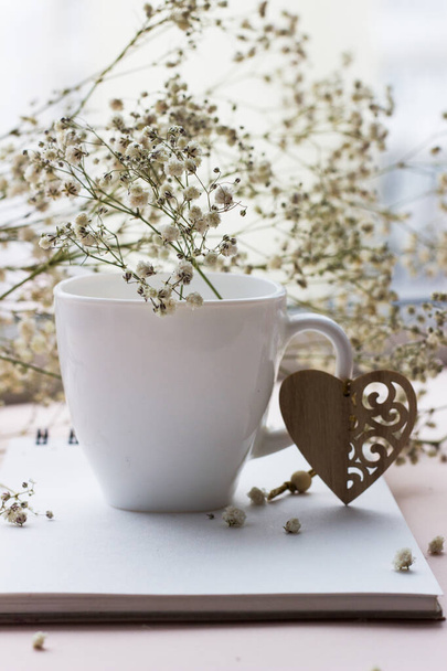 dřevěné srdce a bílý porcelán pohár s kyticí malých bílých květin na světlém pozadí - Fotografie, Obrázek