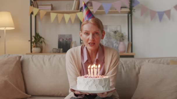 Medio pov disparo de la mujer sonriente en la tapa festiva soplando velas en pastel de cumpleaños teniendo partido en línea sentado en casa en la sala de estar decorada durante coronavirus - Metraje, vídeo