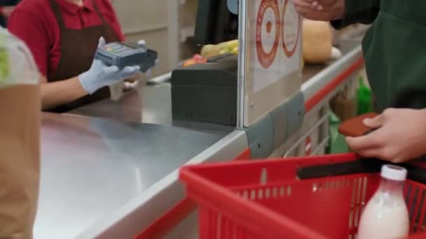 Кассирша в красной форме и защитных перчатках, вручающая клиенту бесконтактную оплату продуктов кредитной картой - Кадры, видео