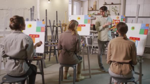 Повний знімок чоловічої художниці, що розмовляє, стоячи перед студентами дорослих жінок у мольбертах у майстер-класі з яскравого мистецтва, що дає майстер-клас з абстрактного живопису
 - Кадри, відео