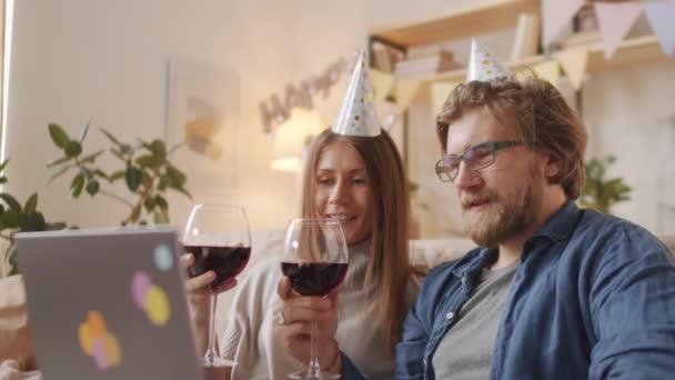 Střední detailní záběr veselého mladého bělošského páru ve slavnostních čepicích cinkajících brýlemi s červeným vínem, zatímco on-line video chat přes notebook vzdáleně slaví narozeniny během coronaviru - Záběry, video