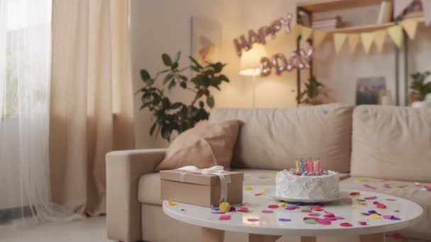 Ninguna persona toma panorámica de acogedora sala de estar decorada con globos de cumpleaños y confeti, pastel de cumpleaños con velas encendidas y caja de regalo artesanal - Imágenes, Vídeo
