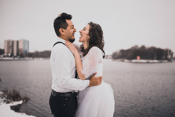 Mexikanische hispanische Männer in Hosenträgern und Frauen küssen sich lachend. Bräutigam und Braut Hochzeitspaar im Winter im Schnee. modisch stilvolle Party-Hochzeitskleidung 2021 - Foto, Bild