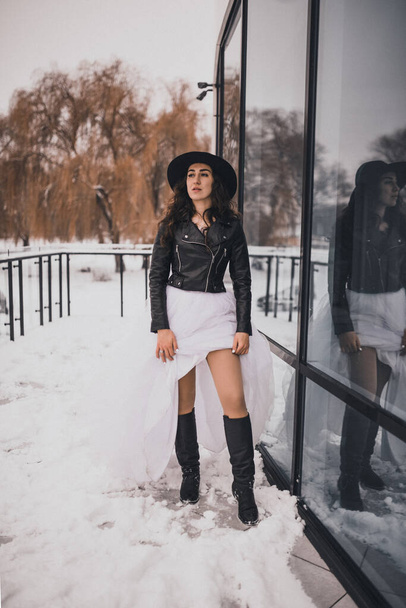 μελαχρινή νύφη γυναίκα σε γάμο λευκό φόρεμα, μαύρο δερμάτινο μπουφάν ποδηλάτης και μαύρο καπέλο σε υψηλές μπότες το χειμώνα στο χιόνι. κομψά ρούχα μόδας κομψό 2021 - Φωτογραφία, εικόνα