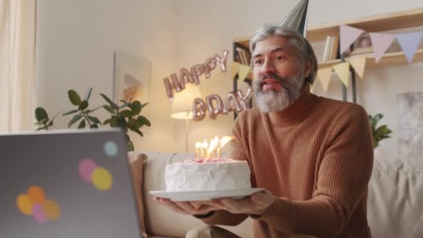 Aufgeweckte Aufnahmen eines kaukasischen Mannes mittleren Alters, der seinen Geburtstag per Laptop feiert und mit seiner Familie per Videoanruf Kerzen auf seiner Geburtstagstorte ausbläst, die allein auf dem Sofa im dekorierten Wohnzimmer sitzt - Filmmaterial, Video