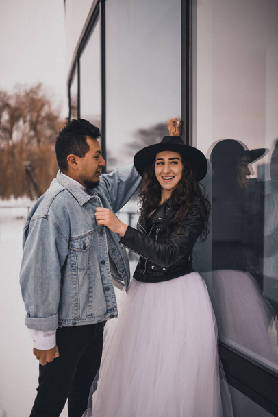 Meksikalı İspanyol adam ve şapkalı kadın sarılıp gülüyorlar. Damat gelinin düğün çifti kışın karlı arka planda vitrinlerin arkasında şık bir parti elbisesi  - Fotoğraf, Görsel