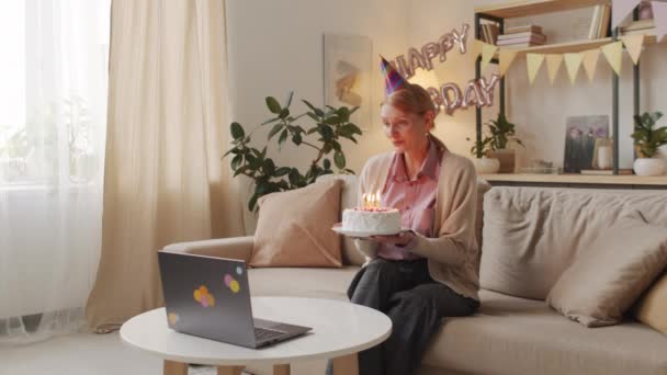 Mediale Aufnahmen einer lächelnden kaukasischen Frau mit Videoanruf am Laptop, die Kerzen auf einer schönen Geburtstagstorte auspustet und während der Sperrung allein zu Hause bleibt - Filmmaterial, Video