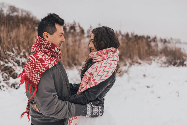νεαρός άνδρας και γυναίκα στέκονται να φιλιούνται και να αγκαλιάζονται. νύφη και γαμπρός ερωτευμένοι ζευγάρι σε πλεκτό μαντήλι και γάντια το χειμώνα με χιόνι και νιφάδες χιονιού. νυφικό. Μεξικανοί - Φωτογραφία, εικόνα