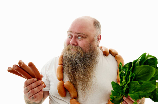 An obese man looking at sausages and holding spinach on the other hand try to choose what is more healthy - Photo, Image