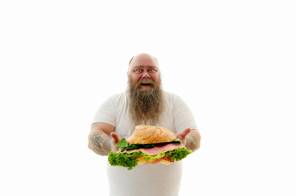 Retrato aislado de un hombre sobrepeso con brazos tatuados sosteniendo una gran hamburguesa con hojas de lechuga y salchichas en rodajas. - Foto, Imagen