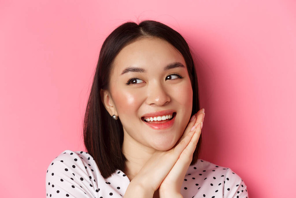 Beauty and skin care concept. Headshot of adorable and dreamy asian woman looking left, smiling and imaging, standing against pink background - Foto, afbeelding
