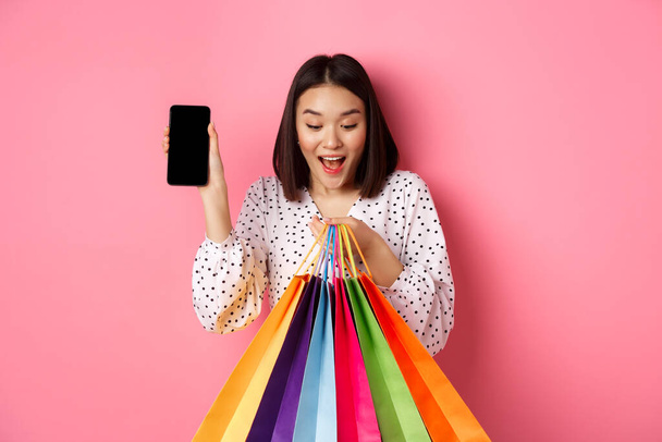Atrakcyjna azjatycka kobieta pokazująca aplikację na smartfona i torby na zakupy, kupująca online za pośrednictwem aplikacji, stojąca na różowym tle - Zdjęcie, obraz