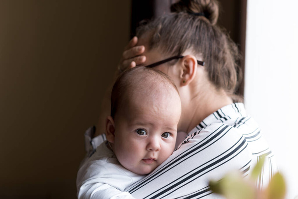 Childhood, motherhood, illness family concepts - upset, tired nervous business woman Lady mother with headache holding infant child newborn baby in her hands stands near window at home indoors - Photo, Image