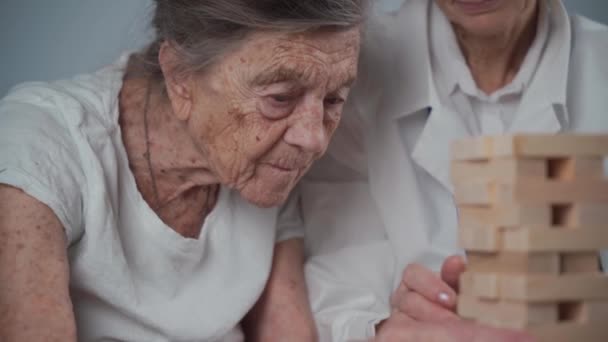Παιχνίδι Τζένγκα. Θέμα είναι η άνοια, η γήρανση και τα παιχνίδια για τους ηλικιωμένους. Καυκάσια ηλικιωμένη γυναίκα χτίζει πύργο από ξύλινα μπλοκ με τη βοήθεια ενός γιατρού ως μέρος ενός παιχνιδιού θεραπείας και Jenga σε ένα σπίτι ασθενών - Πλάνα, βίντεο