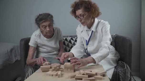 Mature doctor conducting session, therapy for senior patient in nursing home, training fine motor skills for dementia, alzheimer disease and recovery institute by folding wooden blocks, playing - Footage, Video