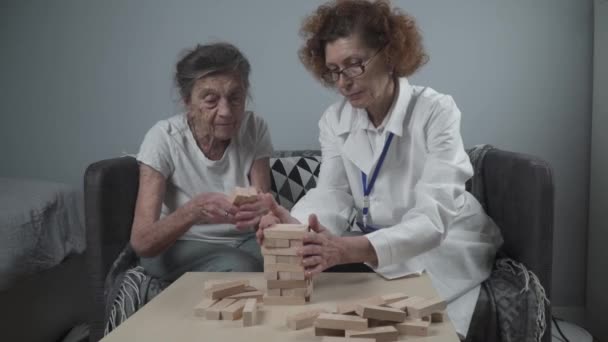 上級女性の練習のスキルは、木製のブロックを構築し、タワーを構築し、それを秋にしないようにしようとすると、 Jengaゲーム。古い患者は、ブロックを引き出します,上の場所,家の中で治療認知症の間にドアをサポート - 映像、動画