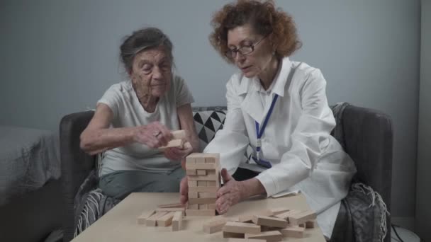 Mature doctor conducting session, therapy for senior patient in nursing home, training fine motor skills for dementia, alzheimer disease and recovery institute by folding wooden blocks, playing jenga - Footage, Video