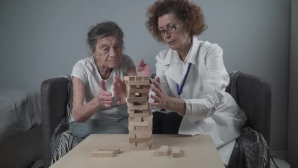 Donna anziana che gioca a Jenga, costruire una torre di blocchi. Medico anziano in camice bianco, che sostiene il paziente anziano, sviluppa la logica, le abilità motorie fini, la malattia di Alzheimer a casa con vecchia donna sola - Filmati, video