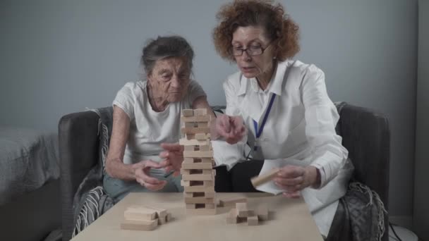 Senior woman playing Jenga, build tower of blocks. Starší lékař v bílém laboratorním plášti, podporující seniorku, rozvíjející se logika, jemné motorické dovednosti, Alzheimerova choroba doma se starou osamělou ženou - Záběry, video