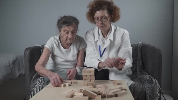 遊び心のある方法で認知症治療は、指や細かい運動能力を訓練し、塔に木製のブロックを構築し、 Jengaを再生します。老人ホームで教育ゲームをプレイするシニア女性90歳と医師 - 映像、動画