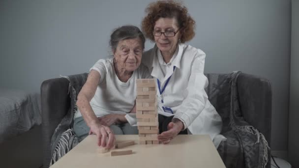 Παιχνίδι Τζένγκα. Θέμα είναι η άνοια, η γήρανση και τα παιχνίδια για τους ηλικιωμένους. Καυκάσια ηλικιωμένη γυναίκα χτίζει πύργο από ξύλινα μπλοκ με τη βοήθεια ενός γιατρού ως μέρος ενός παιχνιδιού θεραπείας και Jenga σε ένα σπίτι ασθενών - Πλάνα, βίντεο