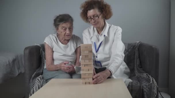 Ανώτερη γυναίκα που παίζει Jenga, χτίζουν πύργο από μπλοκ. Ηλικιωμένος γιατρός σε λευκό εργαστηριακό παλτό, υποστήριξη ανώτερος ασθενής, ανάπτυξη της λογικής, πρόστιμο κινητικές δεξιότητες, Αλτσχάιμερ στο σπίτι με παλιά μοναχική γυναίκα - Πλάνα, βίντεο