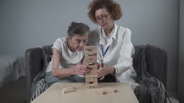 Dementia therapy in playful way, training fingers and fine motor skills, build wooden blocks into tower, playing Jenga. Senior woman 90 years old and doctor playing educational game in nursing home - Footage, Video