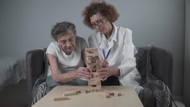 Gioco di Jenga. Tema è la demenza, l'invecchiamento e giochi per gli anziani. Donna anziana caucasica costruisce torre di blocchi di legno con l'aiuto di un medico come parte di una terapia e jenga gioco in una casa di pazienti - Filmati, video