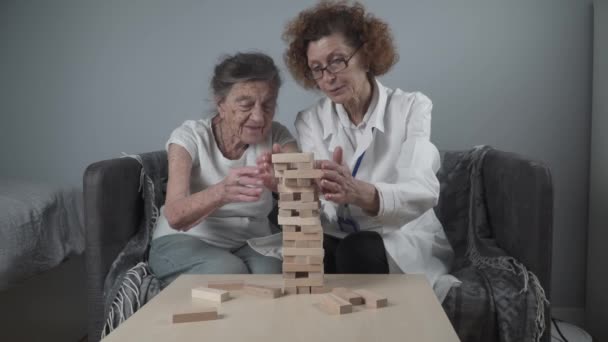 Старшая женщина практиковать навыки построить деревянные блоки, строительство башни и стараясь не дать ему упасть, игра Дженга. Старый пациент вынимает блок, место на вершине, поддержка врача во время терапии слабоумия в доме - Кадры, видео