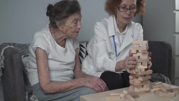 Dementia therapy in playful way, training fingers and fine motor skills, build wooden blocks into tower, playing. Senior woman 90 years old and doctor playing educational game in nursing home - Footage, Video
