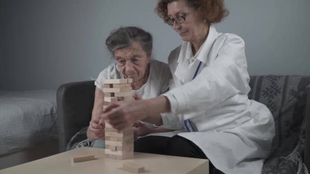 Jogo de Jenga. Tema é demência, envelhecimento e jogos para pessoas idosas. Mulher idosa caucasiana constrói torre de blocos de madeira com a ajuda de um médico como parte de uma terapia e jogo de jenga em uma casa de pacientes - Filmagem, Vídeo