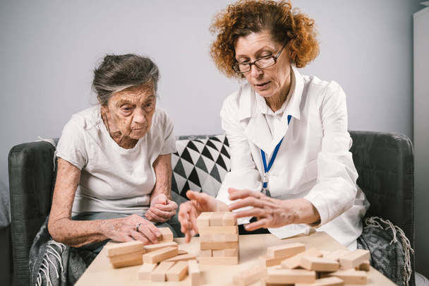シニア女性Jenga再生、ブロックの塔を構築します。高齢者の医師白衣,高齢者をサポート,ロジックを開発,細かい運動能力,高齢者の孤独な女性と自宅でアルツハイマー病. - 写真・画像
