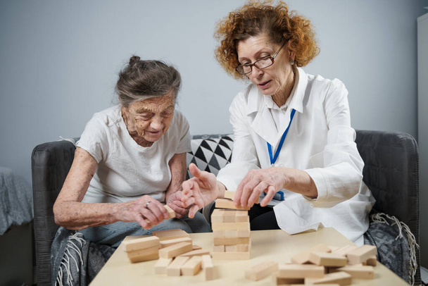 Jenga-peli. Teemana on dementia, ikääntyminen ja pelit vanhuksille. Valkoihoinen vanhempi nainen rakentaa torni puiset lohkot avulla lääkäri osana hoitoa ja jenga peli potilaan kotona. - Valokuva, kuva