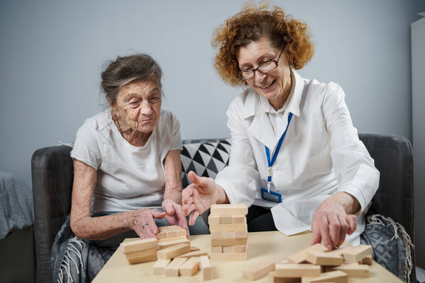 Senior woman practice skills, build wooden blocks, build tower and try not to let it fall, Jenga game. Old patient pull out block, place on top, support doctor during therapy dementia in house. - Photo, Image
