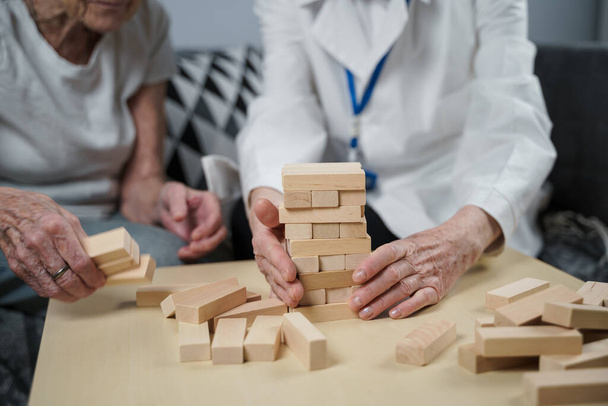 Jenga game. Theme is dementia, aging and games for old people. Caucasian senior woman builds tower of wooden blocks with the help of a doctor as part of a therapy and jenga game at a patient's home. - Photo, Image