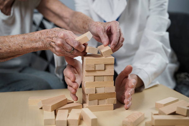 遊び心のある方法で認知症治療は、指や細かい運動能力を訓練し、塔に木製のブロックを構築し、 Jengaを再生します。老人ホームで教育ゲームをプレイするシニア女性90歳と医師. - 写真・画像