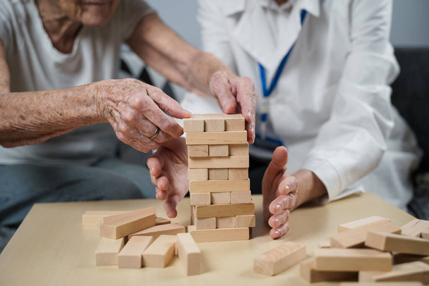 Jenga game. Theme is dementia, aging and games for old people. Caucasian senior woman builds tower of wooden blocks with the help of a doctor as part of a therapy and jenga game at a patient's home. - Foto, imagen
