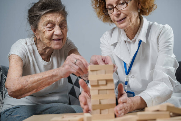 Dementia therapy in playful way, training fingers and fine motor skills, build wooden blocks into tower, playing Jenga. Senior woman 90 years old and doctor playing educational game in nursing home. - Photo, Image