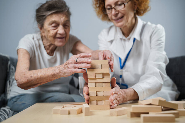Jenga game. Theme is dementia, aging and games for old people. Caucasian senior woman builds tower of wooden blocks with the help of a doctor as part of a therapy and jenga game at a patient's home. - Foto, Bild