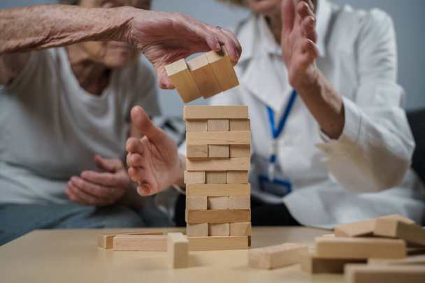 Senior woman practice skills, build wooden blocks, build tower and try not to let it fall, Jenga game. Old patient pull out block, place on top, support doctor during therapy dementia in house. - Photo, Image