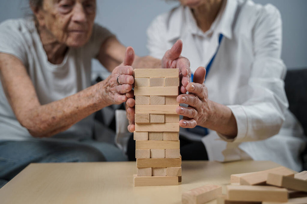 Jeu de Jenga. Le thème est la démence, le vieillissement et les jeux pour les personnes âgées. Femme âgée caucasienne construit tour de blocs de bois avec l'aide d'un médecin dans le cadre d'une thérapie et jenga jeu à la maison d'un patient. - Photo, image