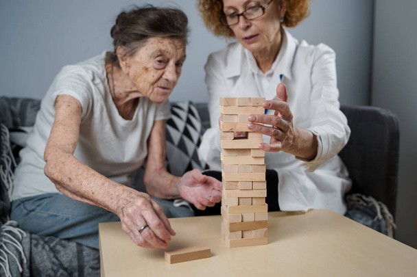 Juego de Jenga. El tema es la demencia, el envejecimiento y los juegos para los ancianos. Mujer mayor caucásica construye torre de bloques de madera con la ayuda de un médico como parte de una terapia y un juego de jenga en el hogar de un paciente. - Foto, Imagen