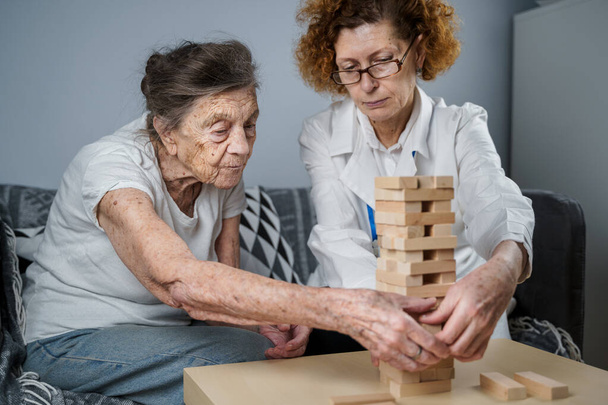 Ανώτερη γυναίκα που παίζει Jenga, χτίζουν πύργο από μπλοκ. Ηλικιωμένος γιατρός σε λευκό εργαστηριακό παλτό, υποστήριξη ανώτερος ασθενής, ανάπτυξη της λογικής, πρόστιμο κινητικές δεξιότητες, Αλτσχάιμερ στο σπίτι με παλιά μοναχική γυναίκα. - Φωτογραφία, εικόνα
