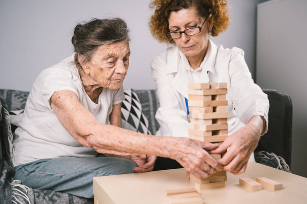 Bunama terapisi, parmaklar ve motor beceriler geliştirmek, kuleye tahta bloklar yapmak, Jenga oynamak. Yaşlı kadın 90 yaşında ve doktor huzurevinde eğitim oyunu oynuyor. - Fotoğraf, Görsel