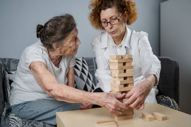 Jenga-Spiel. Themen sind Demenz, Altern und Spiele für alte Menschen. Kaukasische Seniorin baut Turm aus Holzklötzen mit Hilfe eines Arztes im Rahmen eines Therapie- und Jenga-Spiels bei einem Patienten zu Hause. - Foto, Bild