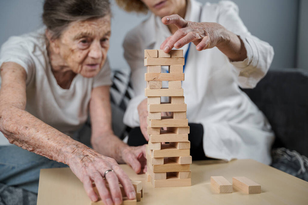 Старша жінка грає в Дженгу, будує башту з блоків. Літній лікар у білій лабораторії, що підтримує старшого пацієнта, розвиває логіку, чудові моторні навички, хворобу Альцгеймера вдома зі старою самотньою жінкою.. - Фото, зображення